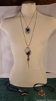 Photo showing four necklaces, two worn on a mannequin and two with black cord chains laid flat on a surface.