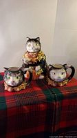 Front view of three Nantucket Pottery ceramic cats: one teapot and two tea cups, sitting on a red and green plaid cloth.