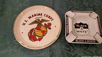 Both ash trays side by side on a green patterned cloth background, left is Marine Corps ash tray, right is Black & White ash tray.