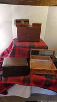 Five wooden jewelry boxes displayed on a red and green plaid cloth. Includes a large chest-style box with drawers, small simple wood box, rectangular box with interior mirror, an Asian-painted box with metal latch, and a small thin box.