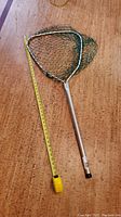 Fishing net with triangular frame, green mesh netting, and aluminum handle next to a yellow measuring tape showing the length.