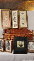 Large open wicker basket with lid and carrying handles holding several pieces of framed artwork, including oval bird and floral illustrations, and a black framed nature image. Two small framed floral artworks placed in front of basket.