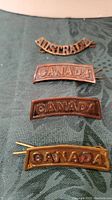 Four military badges on dark fabric background, including one 'AUSTRALIA' and three 'CANADA' badges.