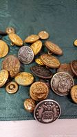Various military buttons in different sizes and metal tones scattered on a green cloth background, showing detailed embossed designs such as eagles, shields, and anchors.