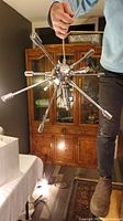 Photo showing a person holding the spudnik hanging ceiling light with multiple chrome rods extending from the spherical center, set against a background with wooden furniture.
