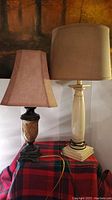 Pair of two table lamps placed on a patterned red and black fabric surface against a wall with brown artwork behind. The lamps have different bases and shades. The cream colored lamp base supports a fabric cylindrical beige shade. The darker decorative base lamp supports a smaller tapered rectangular brown shade with ripple inside.