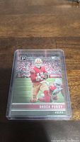 Single photo of a Brock Purdy football rookie card in a clear protective case showing the player in red and gold San Francisco 49ers uniform on the field.