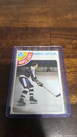 Photo shows a Darryl Sittler hockey trading card in a protective plastic case on a wooden surface. The card shows Sittler in his Maple Leafs uniform, ready to play.