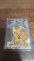 Front view of Kobe Bryant rookie card in protective plastic case showing the player in Lakers yellow jersey number 8 with NBA Topps branding and Lakers logo.