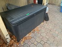 Front view of large black outdoor storage bin with woven wicker-like texture, showing curved lid and lockable latch.
