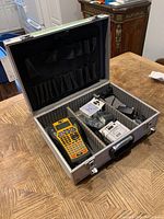 Open Plano aluminum hard case on wooden table showing label printer and accessories