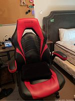 Gaming chair in black and red vinyl upholstery with branding on headrest and padded armrests, near bed.