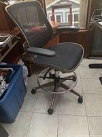 Office chair with black mesh back and seat, armrests, metallic base with caster wheels and chrome ring footrest, positioned on a tiled floor next to window and plastic storage bin.