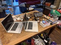 Two MacBook Pro A1297 laptops side by side on wood table with cables, power adapters, power strip, game discs and controllers visible