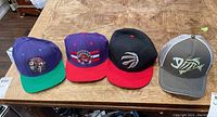 Four snapback caps displayed on a wooden table showing two Raptors hats, one skateboard-themed, and one fishing-themed hat