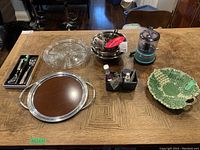 Photo shows entire lot on a wood table: KitchenAid mini food processor, stainless steel serving tray, wire rack, stainless steel bowls, ceramic cabbage leaf plate, kitchen utensils with knives, and salad utensils.