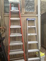 Two ladders side by side against a beige wall: an orange-sided 8-foot ladder and a smaller aluminum ladder with blue top section.