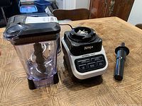 Photo showing Ninja blender base, blending pitcher with lid, and tamper tool on wooden table.