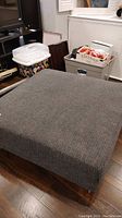 Front angled view of grey upholstered ottoman on wooden floor with household items nearby.