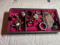 Box with divided compartments containing a variety of metal rings, some with stones and varied designs.