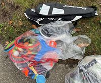 Wide shot of plastic bag containing assorted sports toys including colorful frisbees, ball bats, and other small sports gear outside on pavement and grass.