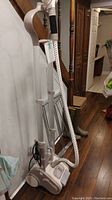 Rowenta clothes steamer standing upright against wall near stairs, beige/white body, wrapped steam hose and a handle with a fabric cover.