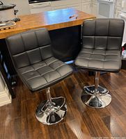 Two black vinyl bar stools with chrome metal bases and adjustable height, placed next to a wooden kitchen island.