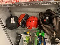 Three helmets placed on metal shelving with bracket support, including black, red, and orange helmets, next to a black tote bag containing shoes.