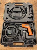 Full view of the SecurityMan tool camera kit opened in molded black plastic carrying case showing orange and black handheld main unit with connected flexible camera cable, display monitor, power adapter, and cables.