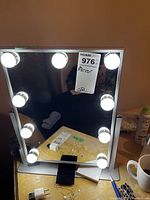 Square mirror with eight bulbs lit around the frame on a table with various items visible around it.