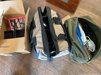 Three items shown: a cardboard box containing a bike hanger and lock, a beige and blue soft tool bag, and a green soft tool bag with tools inside.