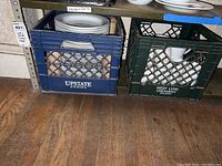 Crate and Barrel white ceramic plates packed in a blue milk crate from Upstate Farms and mugs packed in a green crate from West Lynn Creamery on a wooden floor shelf.
