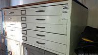 Angle view of metal flat file cabinet with label holders and drawer handles, visible scuffs and scratches on the surface.