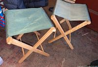 Two light blue canvas-seated wooden folding stools side by side, showing general wear on wood and fabric.