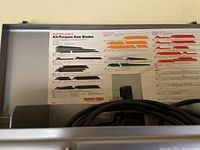 View of the inside saw blade guide chart, showing types of all-purpose saw blades by color and cutting purpose, inside the metal storage case with power cord visible.