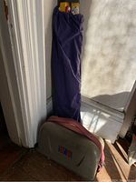 Tripod in purple fabric bag standing upright next to a hard carrying case with red strap.