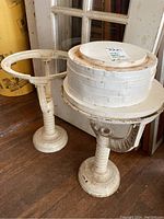 Two white vintage metal table bases on a wooden floor. One base holds multiple round wooden table tops stacked.