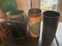 Four vintage metal waste baskets displayed on a table near window, showing overall designs and condition.