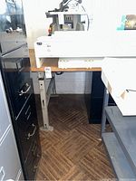 Front angle showing the wood top and adjustable metal legs of the work bench, positioned between black filing cabinets.