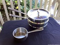All Clad cookware set with double boiler, insert strainer, glass lid, and small cup pot shown from side angle on black cloth.