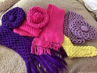 Various wool hats, mittens and scarves in bright colors including hot pink, lavender, purple and yellow, arranged on a blanket.