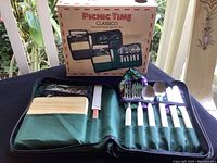Photo showing the full picnic set with the box, green travel pack, wooden cutting board, cheese spreader, and cutlery with white handles including forks, spoons, and knives laid out.