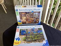Lot shows two Ravensburger puzzles, one 500-piece and one 300-piece, stacked on a table outdoors.