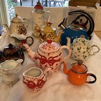 Wide view of the teapots, creamers, and kettle on a white cloth showing overall lot content.