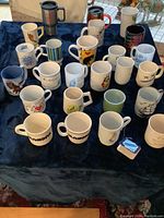 Overview of the 23 coffee cups and 2 to-go cups laid out on dark fabric showing various designs and colors.
