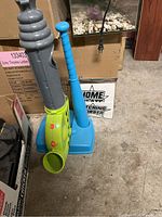 Photo of plastic baseball pitching machine with blue bat and base in a storage area floor.