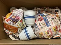 Box containing ceramic mugs wrapped in newspaper, showing two white mugs with blue stripes