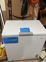 White Midea chest freezer in garage setting with some dirt and storage items around it.