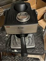 Front view of Mr. Coffee espresso machine showing the stainless steel front panel, black plastic top with a large round knob, and portafilter attached. The glass coffee pot sits underneath on the drip tray, missing its handle.