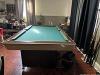 Full view of the pool table showing the green felt playing surface with its wooden frame and legs, indoors next to a window and curtains. The table is surrounded by various household items and cue sticks leaning against the wall.